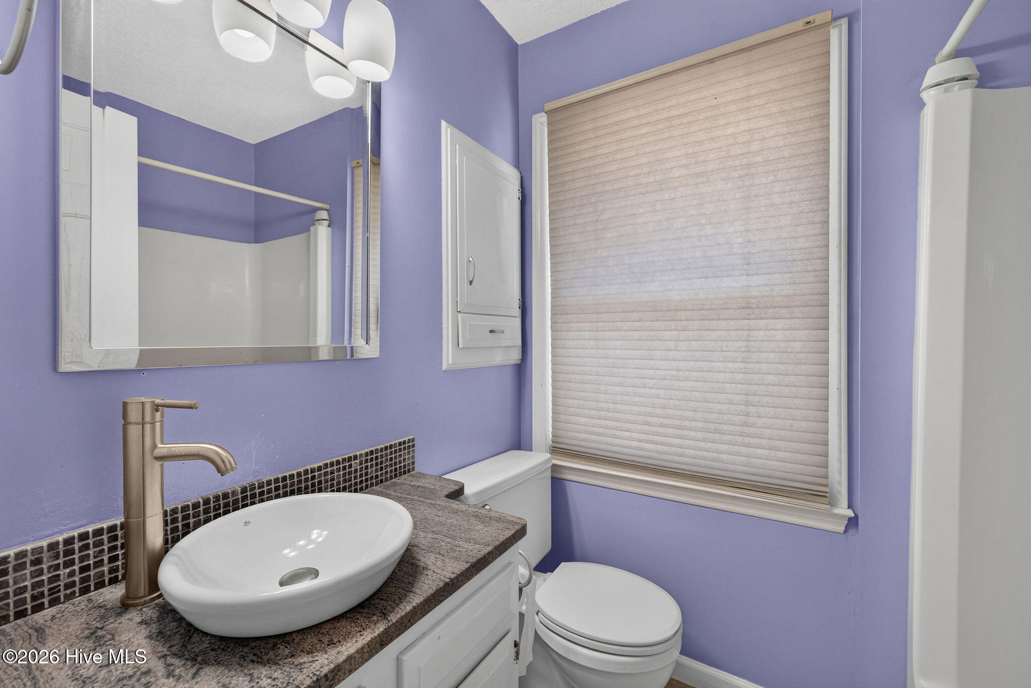 Bathroom with lavender walls, vessel sink, dark mosaic backsplash, and closed window shade.