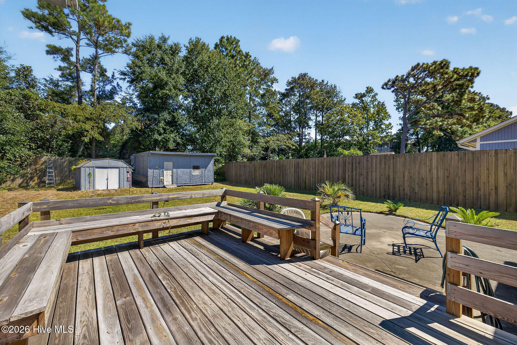 Wooden backyard deck with built-in benches overlooks a fenced yard, storage sheds, and a patio with blue chairs.