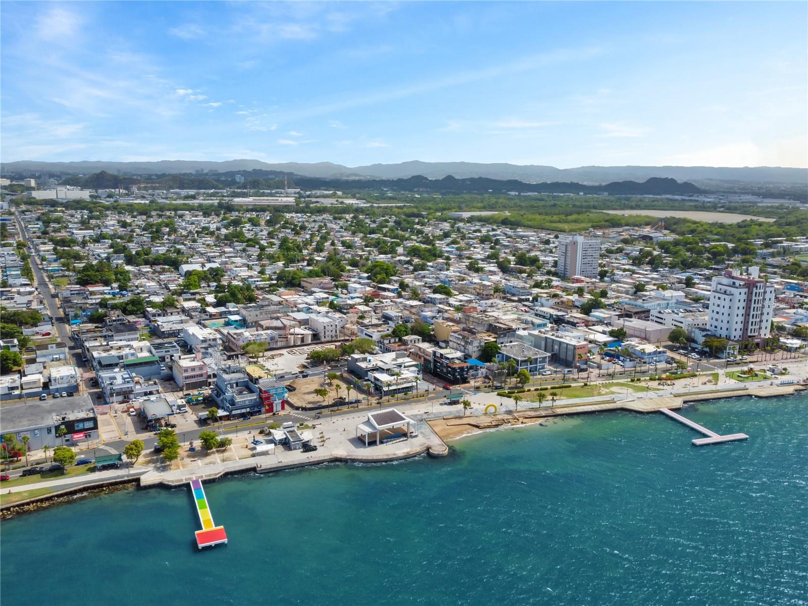 Aerial view of Cataño waterfront, with a colorful pier, dense buildings, and distant hills.