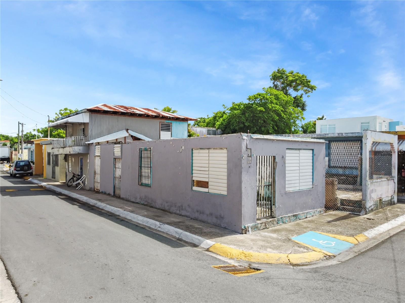 Street-corner view of a low-rise gray property with barred windows and a rusted metal roof.