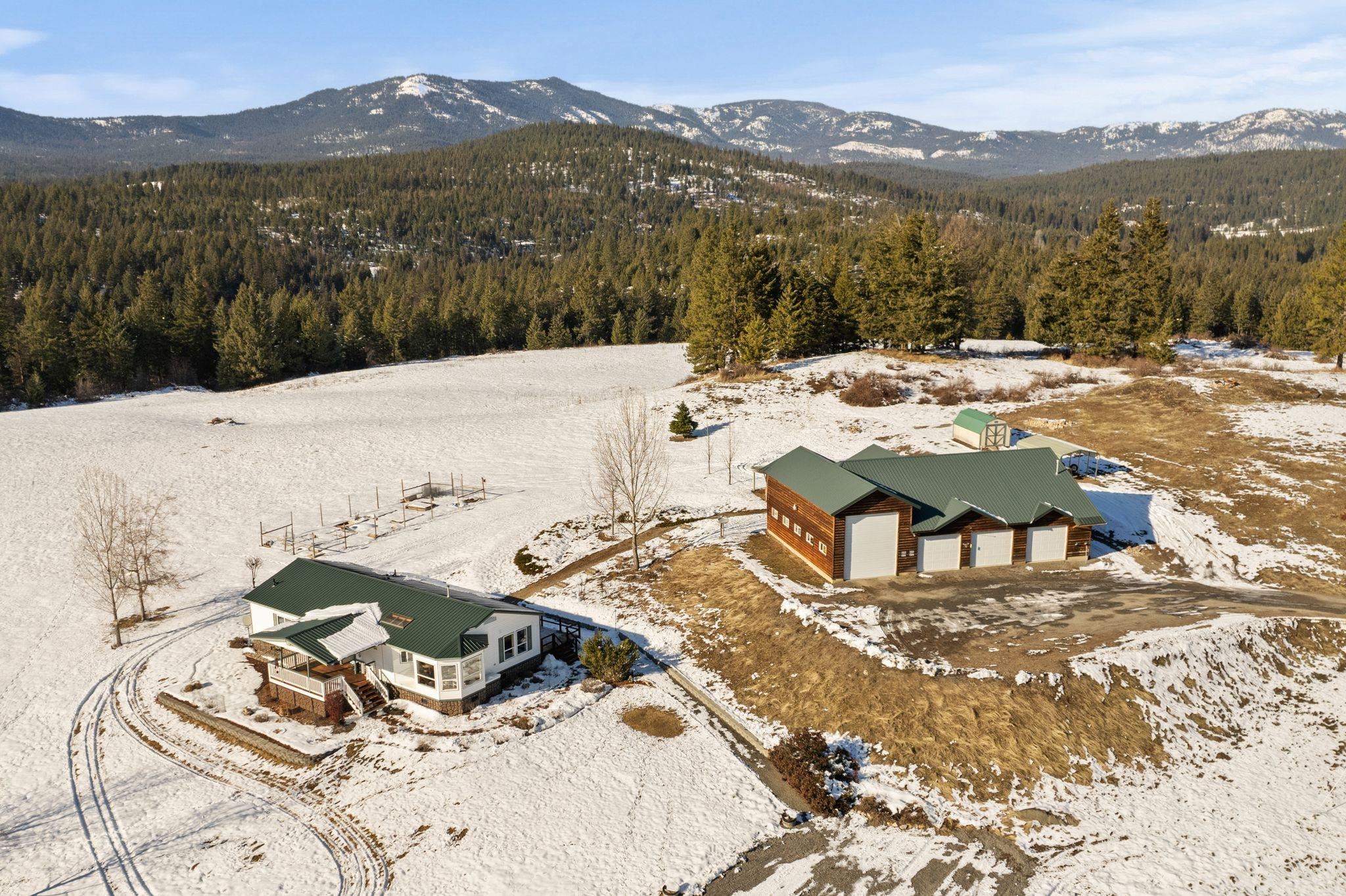 Aerial view of a rural home and large workshop surrounded by snowy fields, evergreen forest, and mountains.
