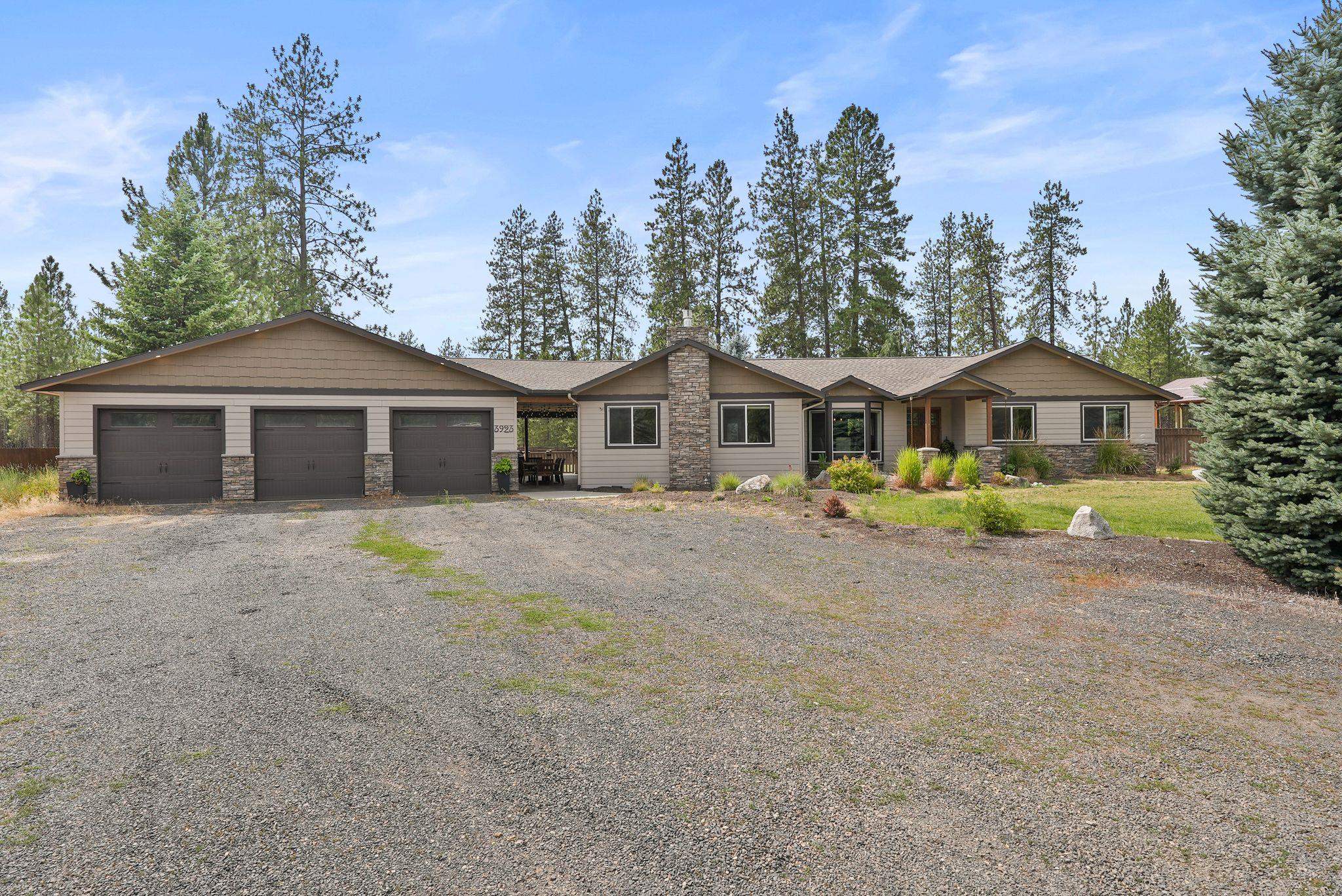 Single-story home with three-car garage, stone accents, broad gravel driveway, and pine trees.
