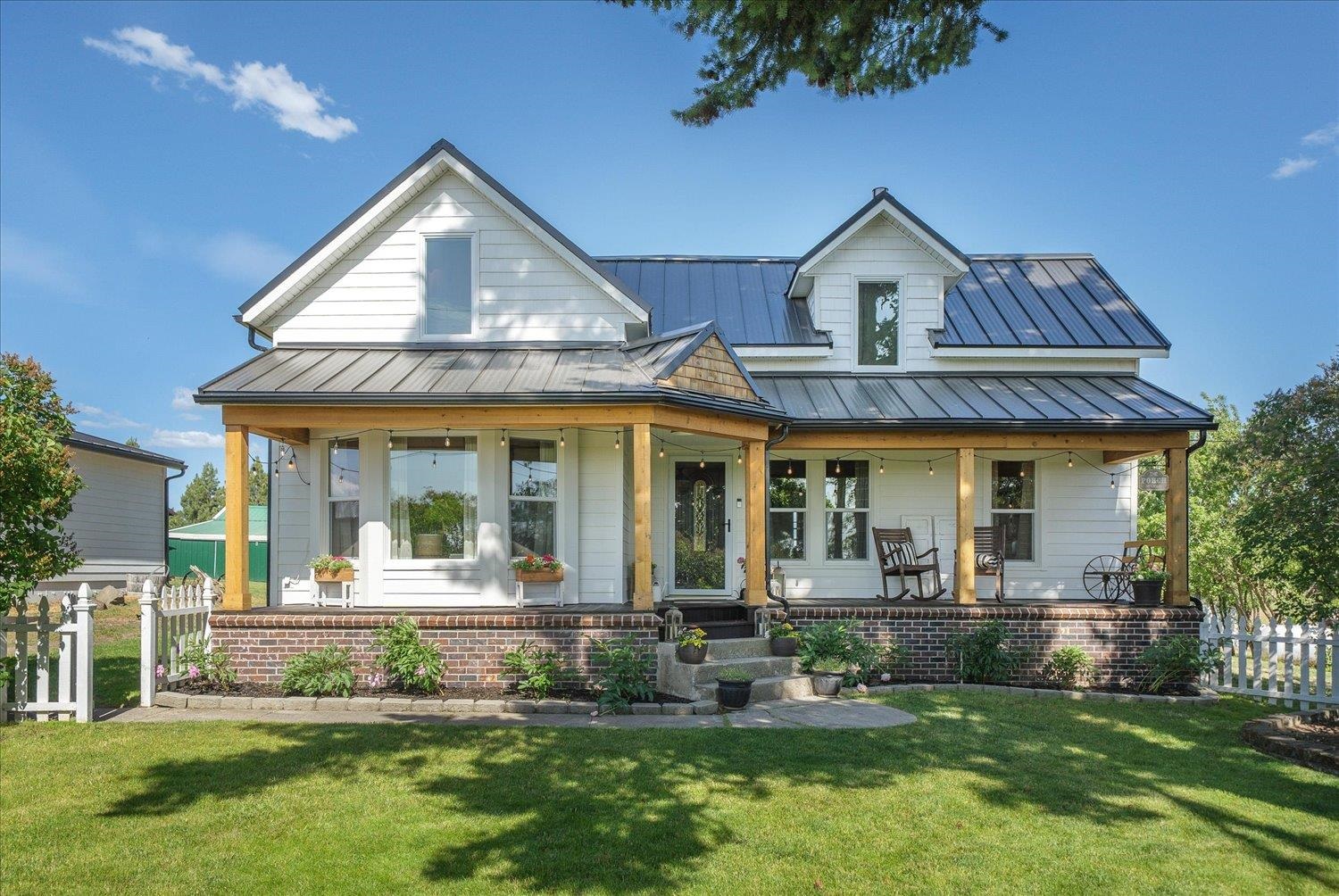 White farmhouse with a metal roof, covered porch, rocking chairs, and white picket fence.