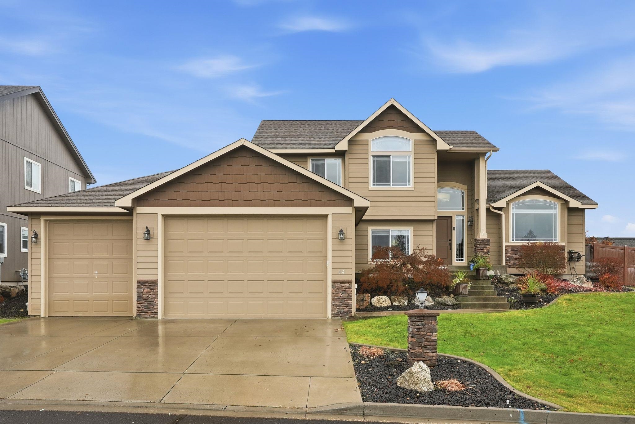 Two-story tan house with three garage doors, stone accents, and a landscaped front yard.
