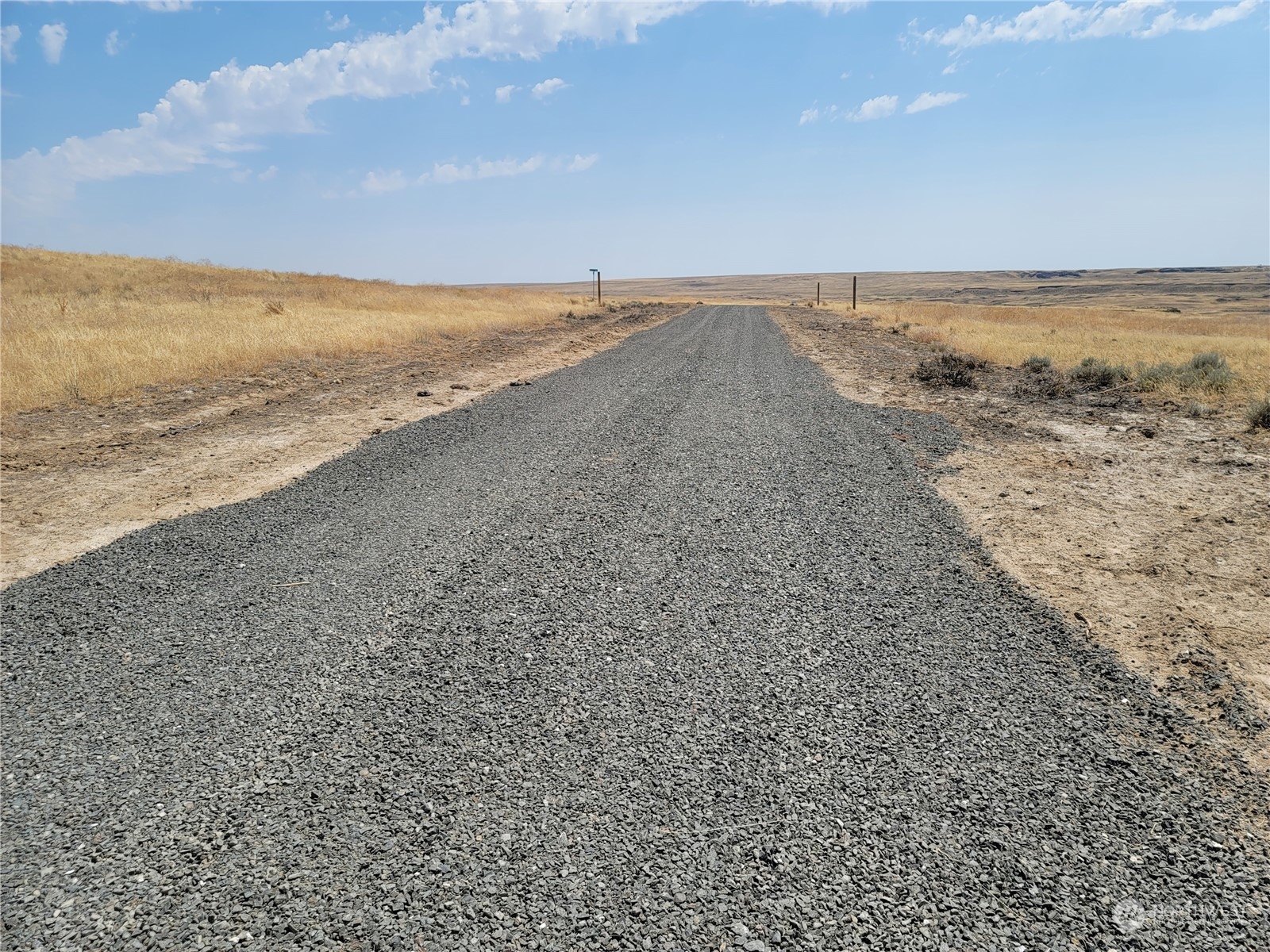 Gravel access road through open, dry grassland beneath a partly cloudy sky.
