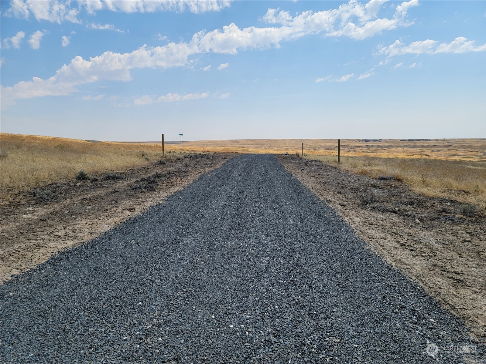 Gravel access road through open grassland, with fence posts and rolling hills in the distance.