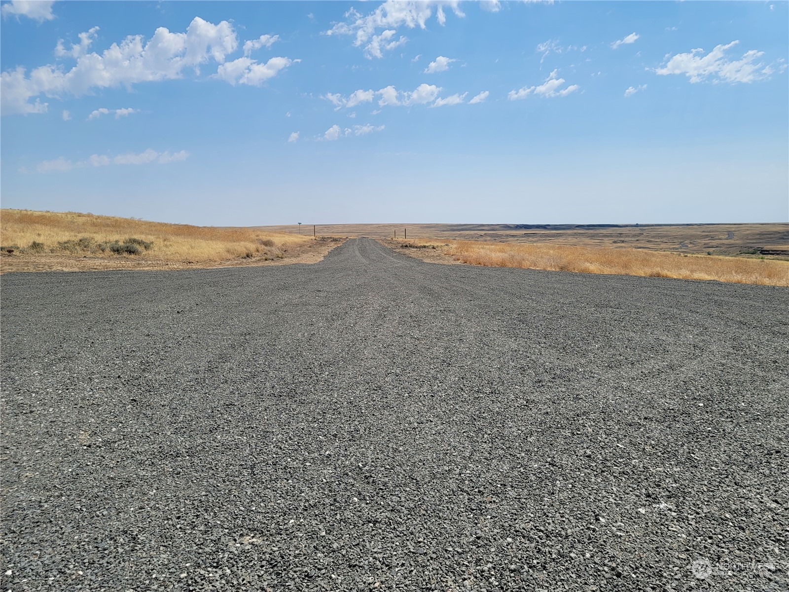 Gravel road through open, rolling grassland beneath a blue sky.