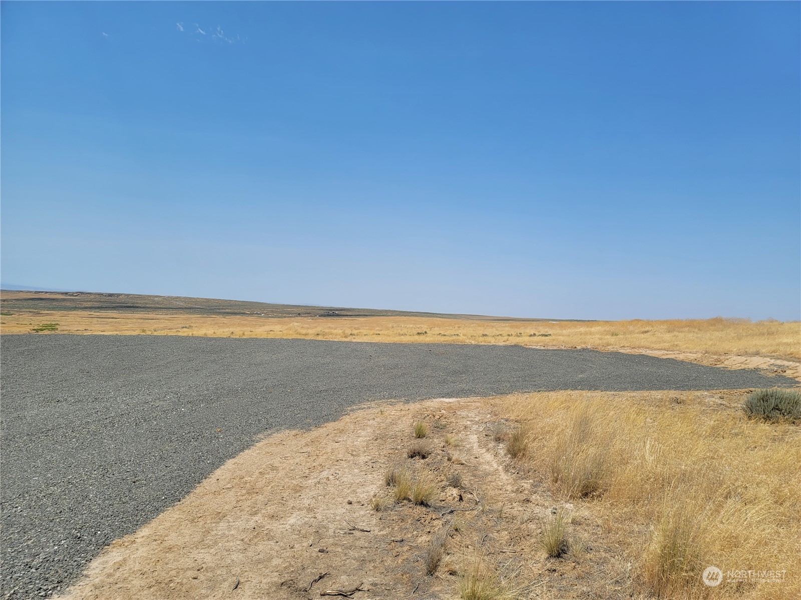 Vacant rural lot with a curving gravel road through dry grassland beneath a clear blue sky.