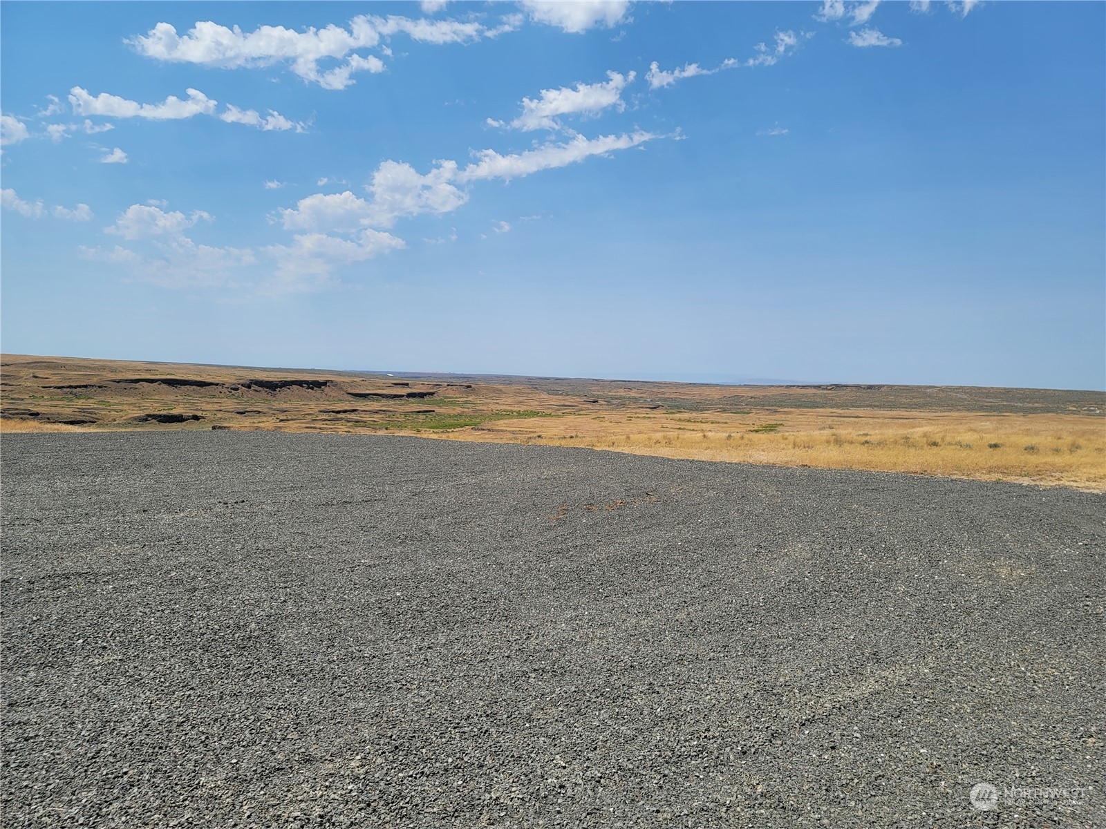 Open vacant land with a broad gravel foreground, rolling grassland, and expansive blue sky.