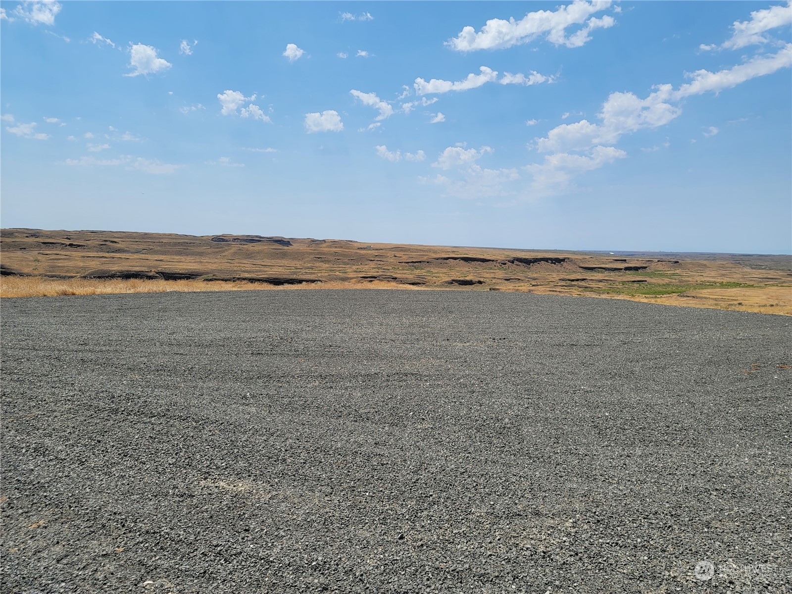 Gravel-covered lot overlooking open, rolling grassland and low rocky ridges beneath a blue sky.