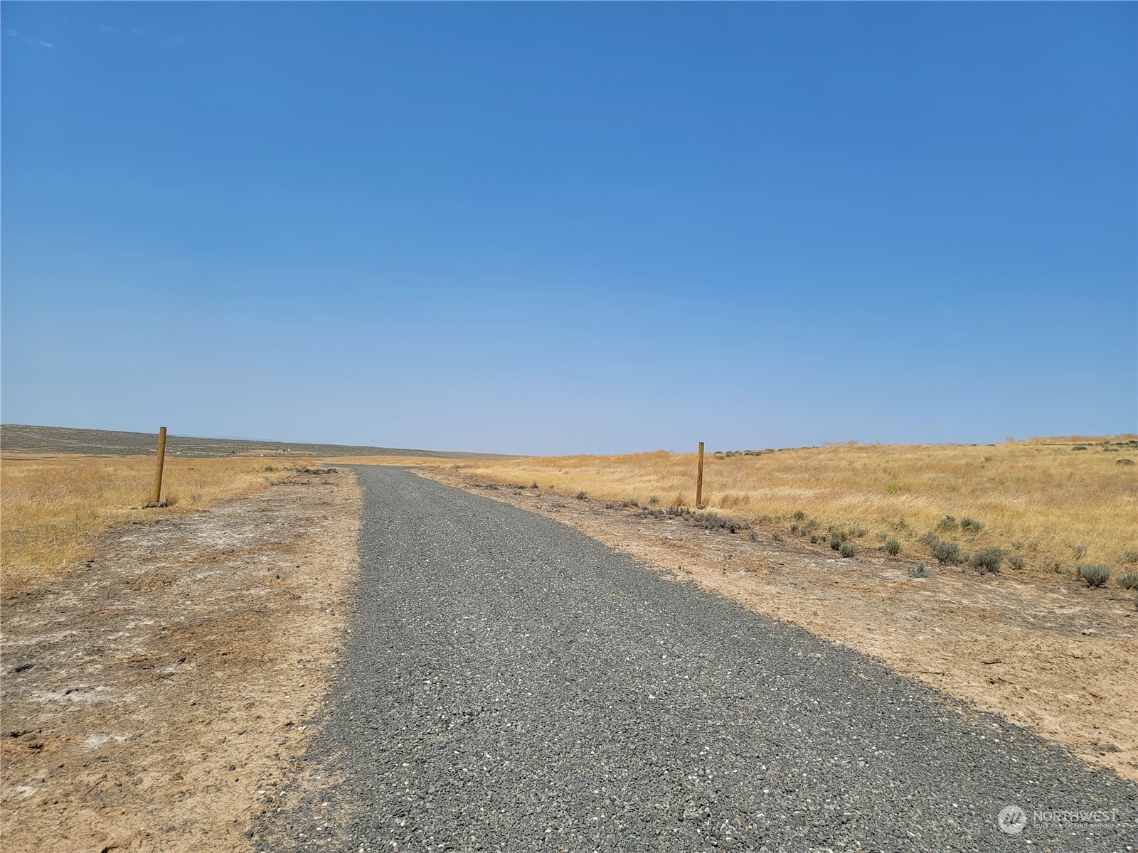 Gravel road through open dry grassland, marked by two fence posts beneath a wide blue sky.