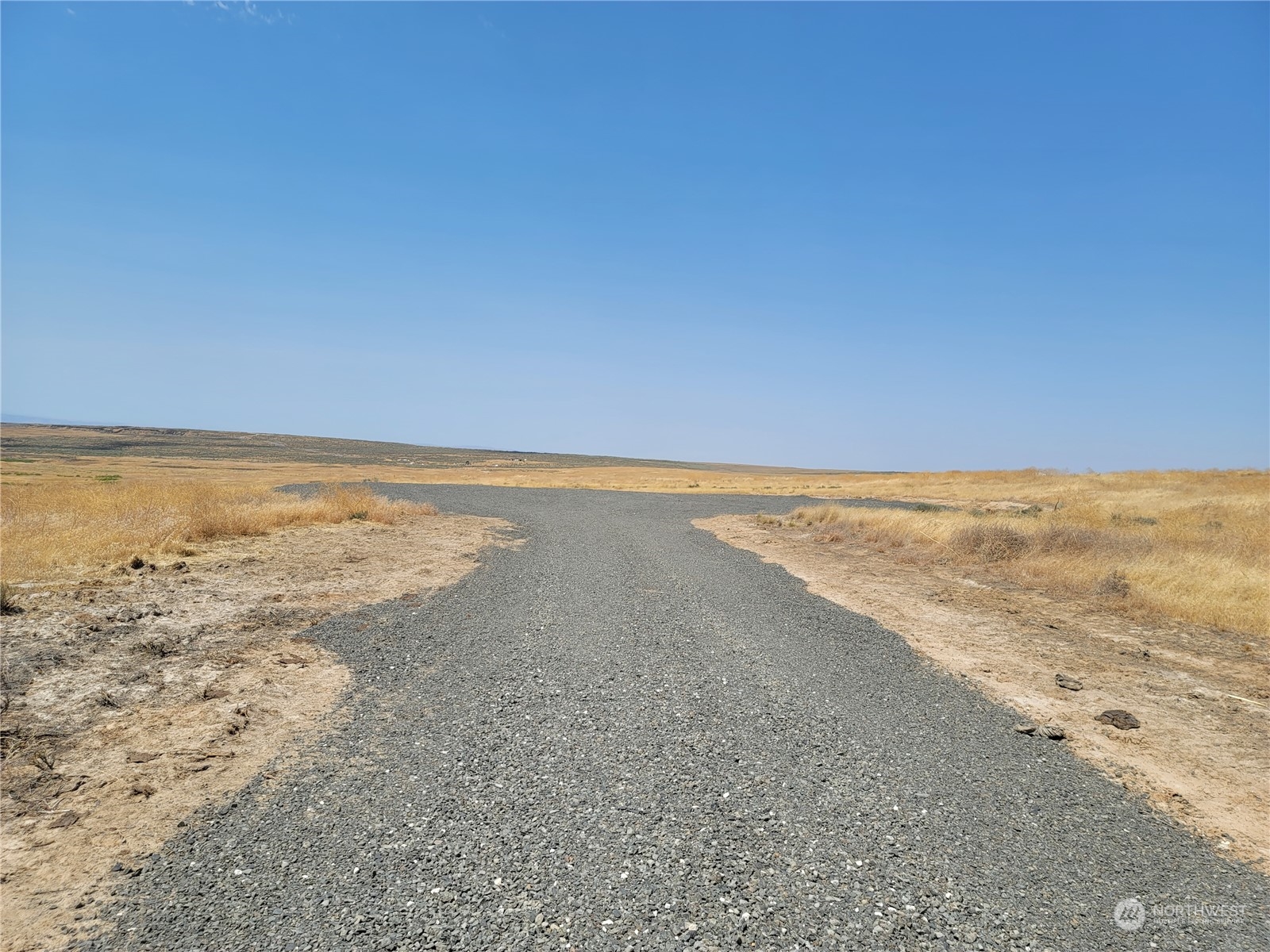 Gravel road through an open rural lot with dry grass and low rolling terrain.