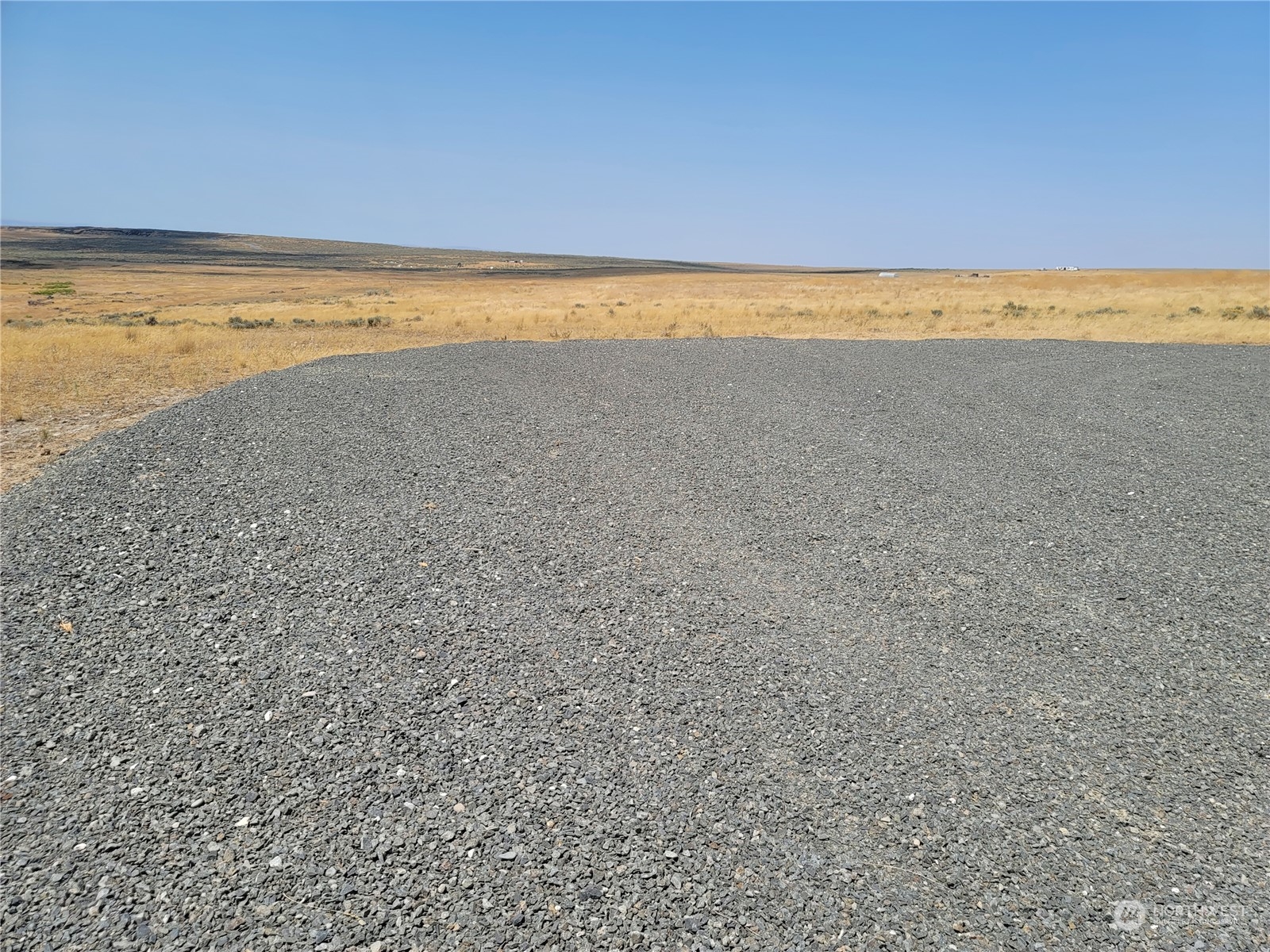Gravel pad on an open rural lot, bordered by dry grassland and distant low hills.