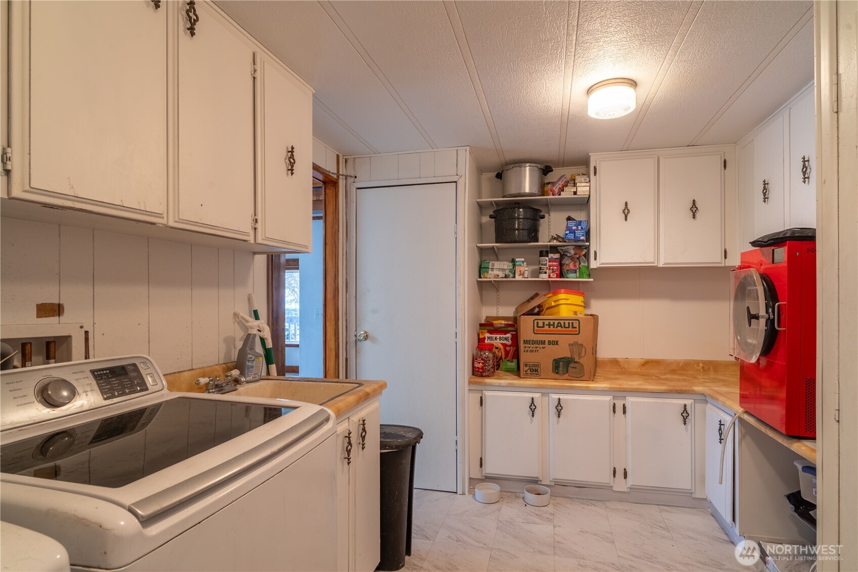 Utility room with a top-loading washer, white cabinets, stocked shelves, and a red machine.