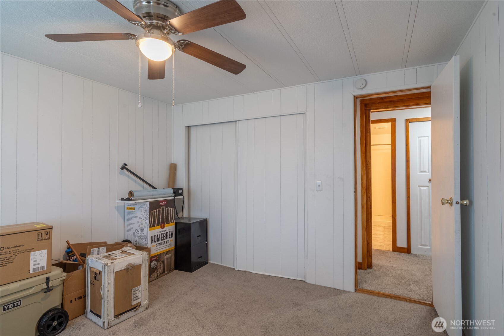 Carpeted room with white paneled walls, a ceiling fan, sliding closet doors, and boxes.