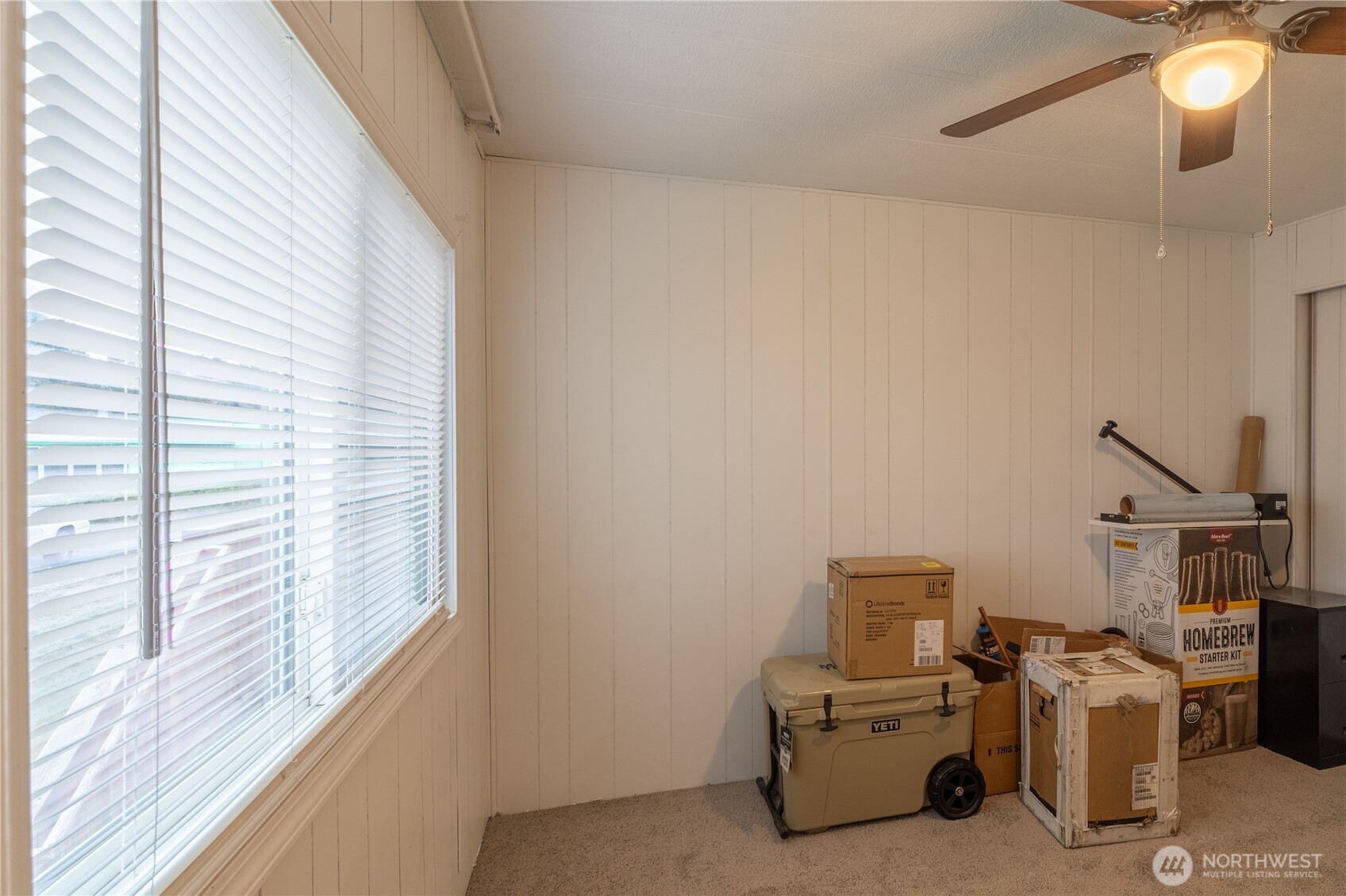 Carpeted room with white blinds, a ceiling fan, and stored boxes beside a wheeled cooler.