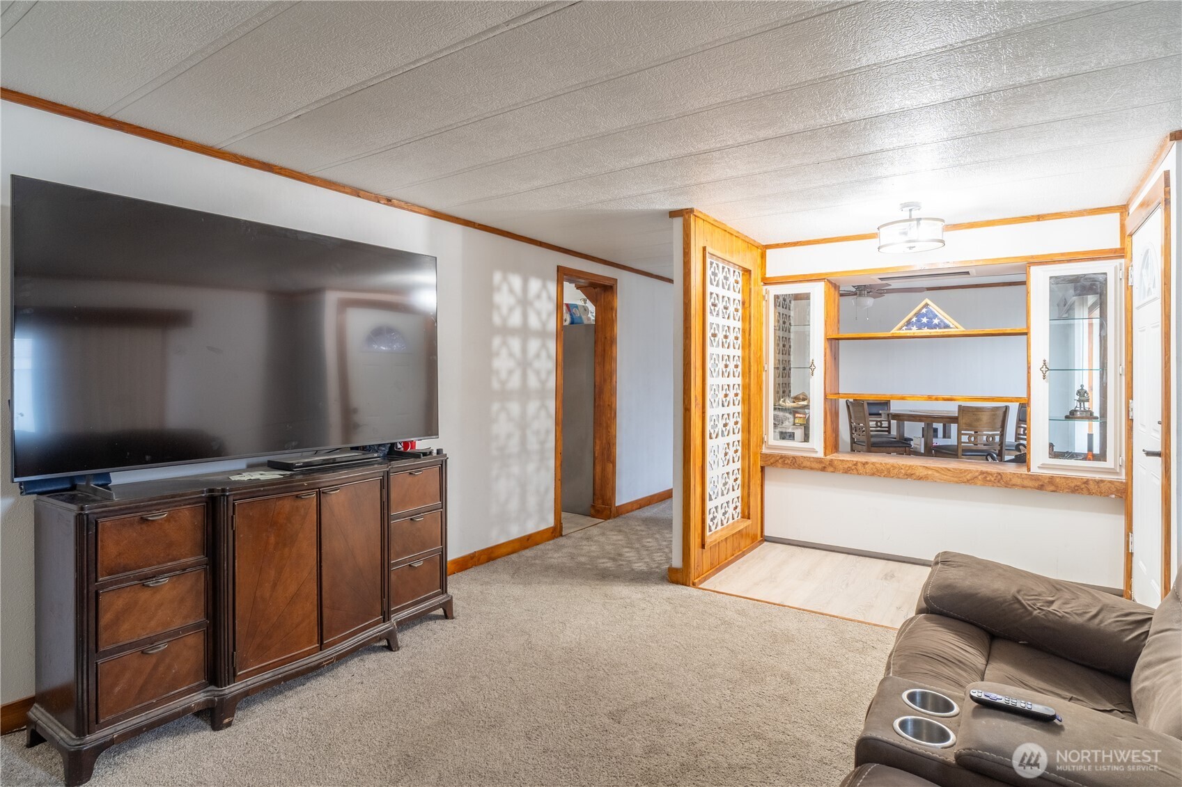 Living room with a large television, beige carpet, and a wood-trimmed pass-through to the dining area.