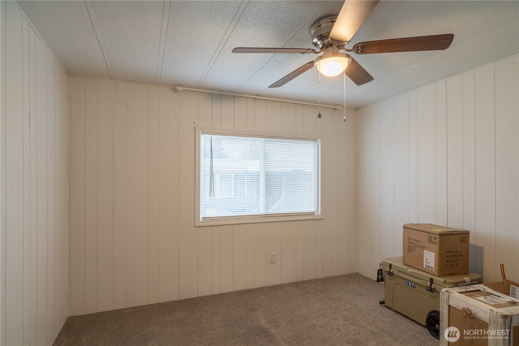 Carpeted room with a window, ceiling fan, and boxes and cooler along one wall.