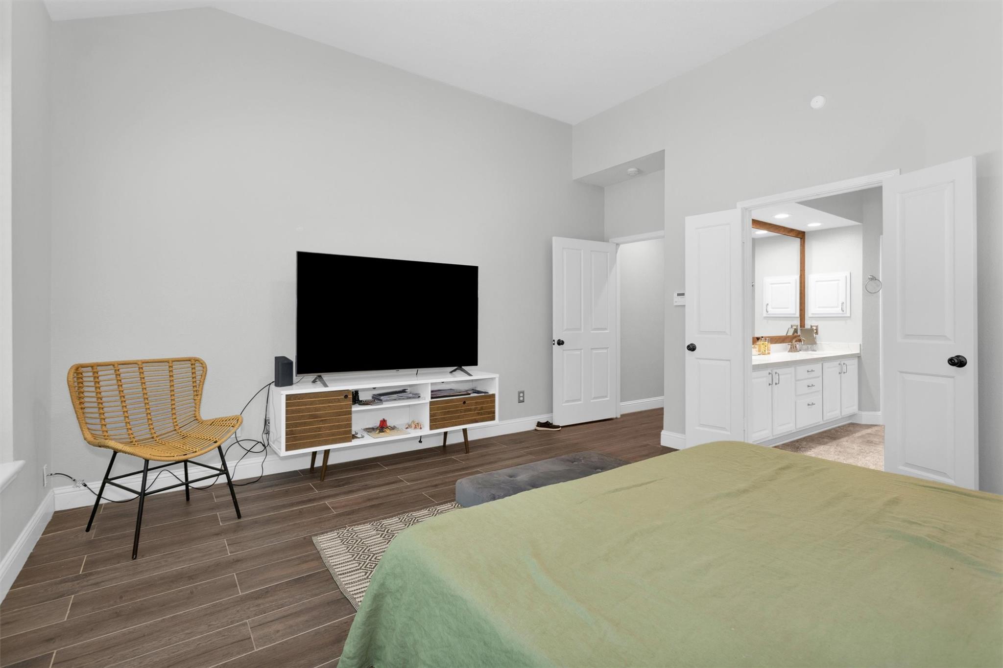 Bedroom with a green bedspread, woven chair, television, and open doors to a bathroom vanity.