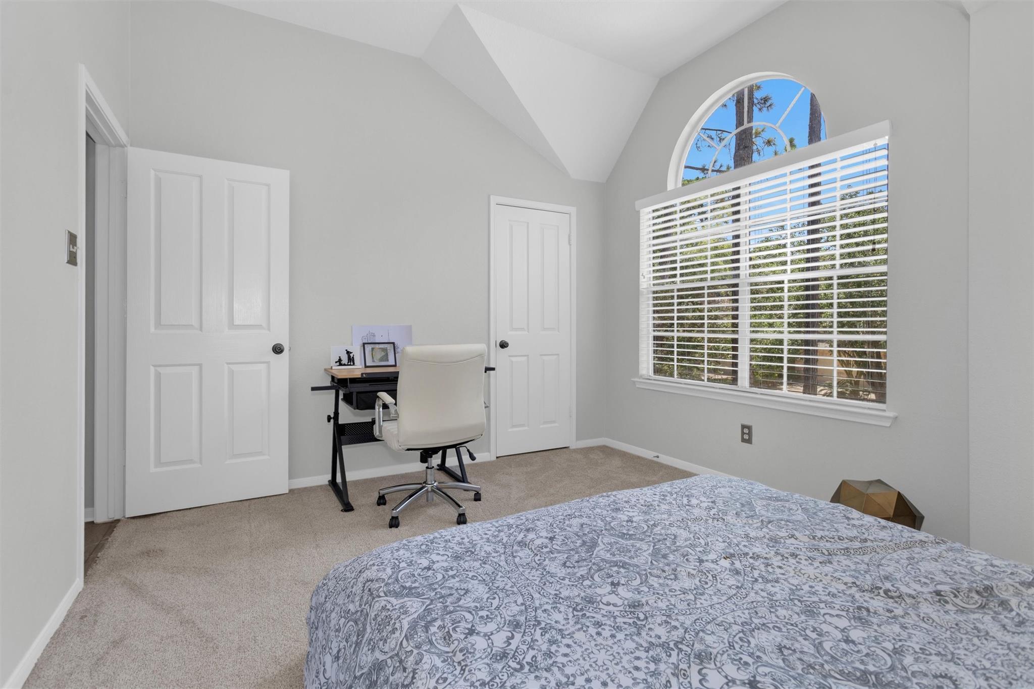 Bedroom with a patterned bedspread, desk and chair, and a large arched window overlooking trees.