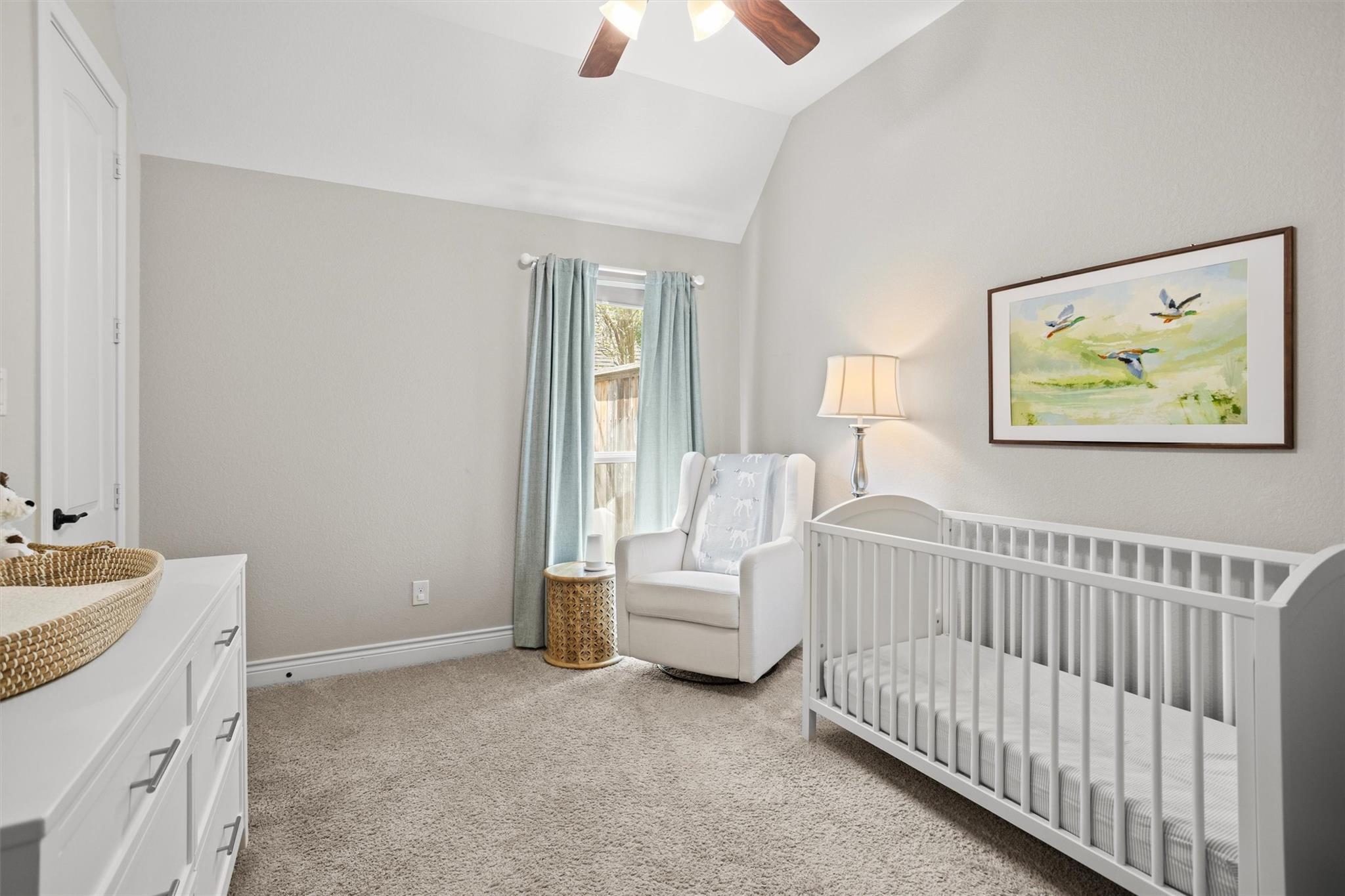 Furnished nursery with a white crib, glider chair, and pale blue curtains beneath a vaulted ceiling.