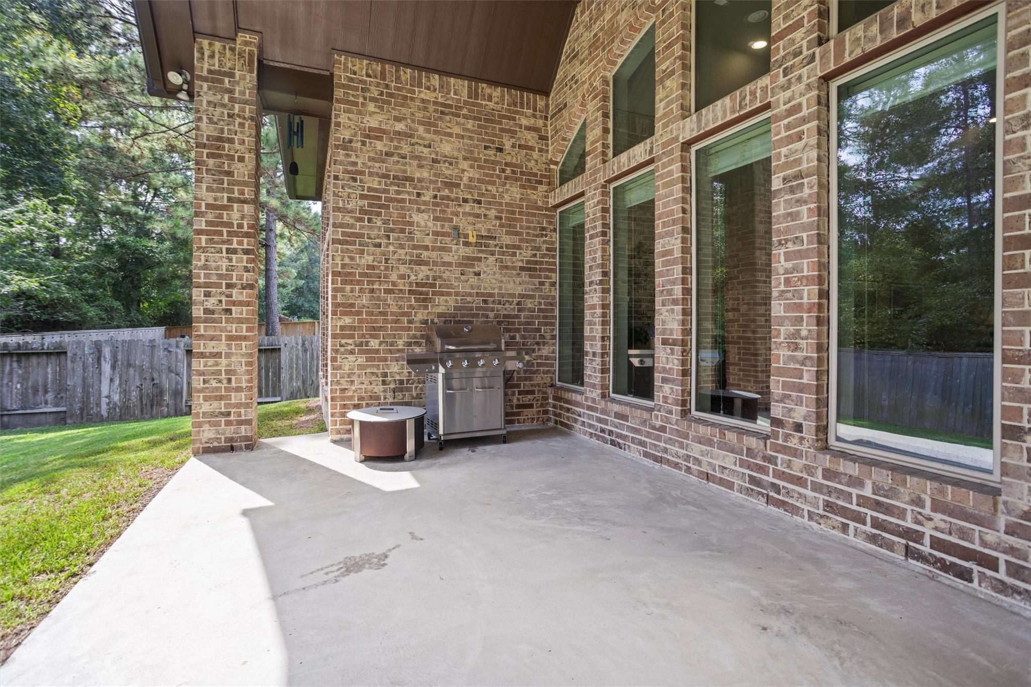 Covered brick patio with a stainless steel grill, tall windows, and views of the fenced yard.