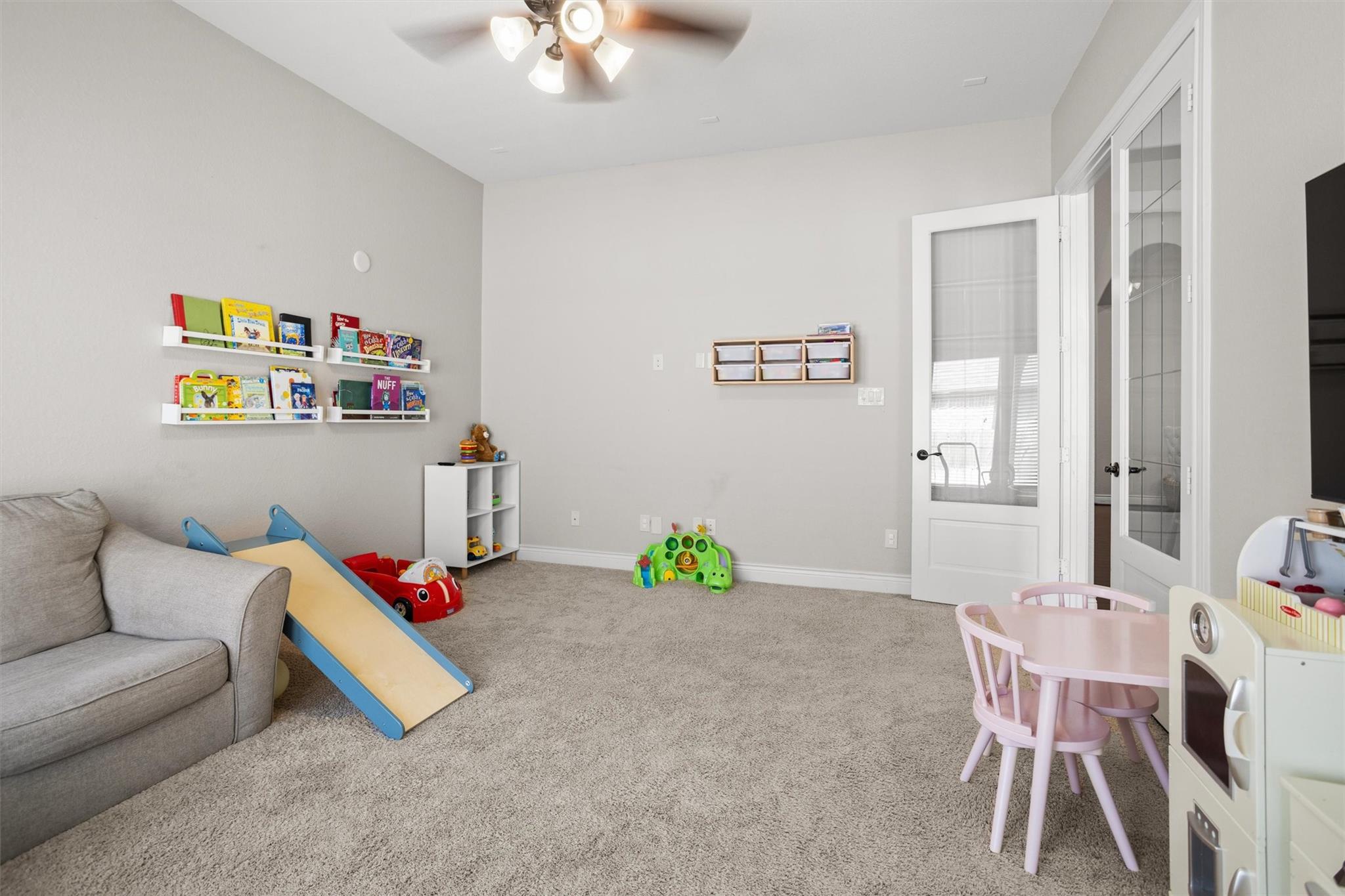 Playroom with a small slide, children’s books on wall shelves, toys, and a pink table and chairs.