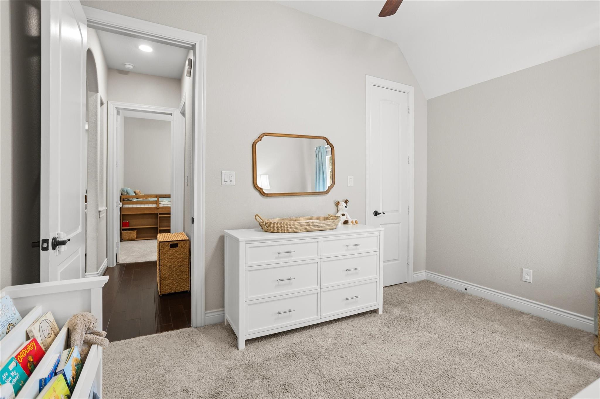 Carpeted nursery with a white dresser, framed mirror, and doorway to a small wooden bed.