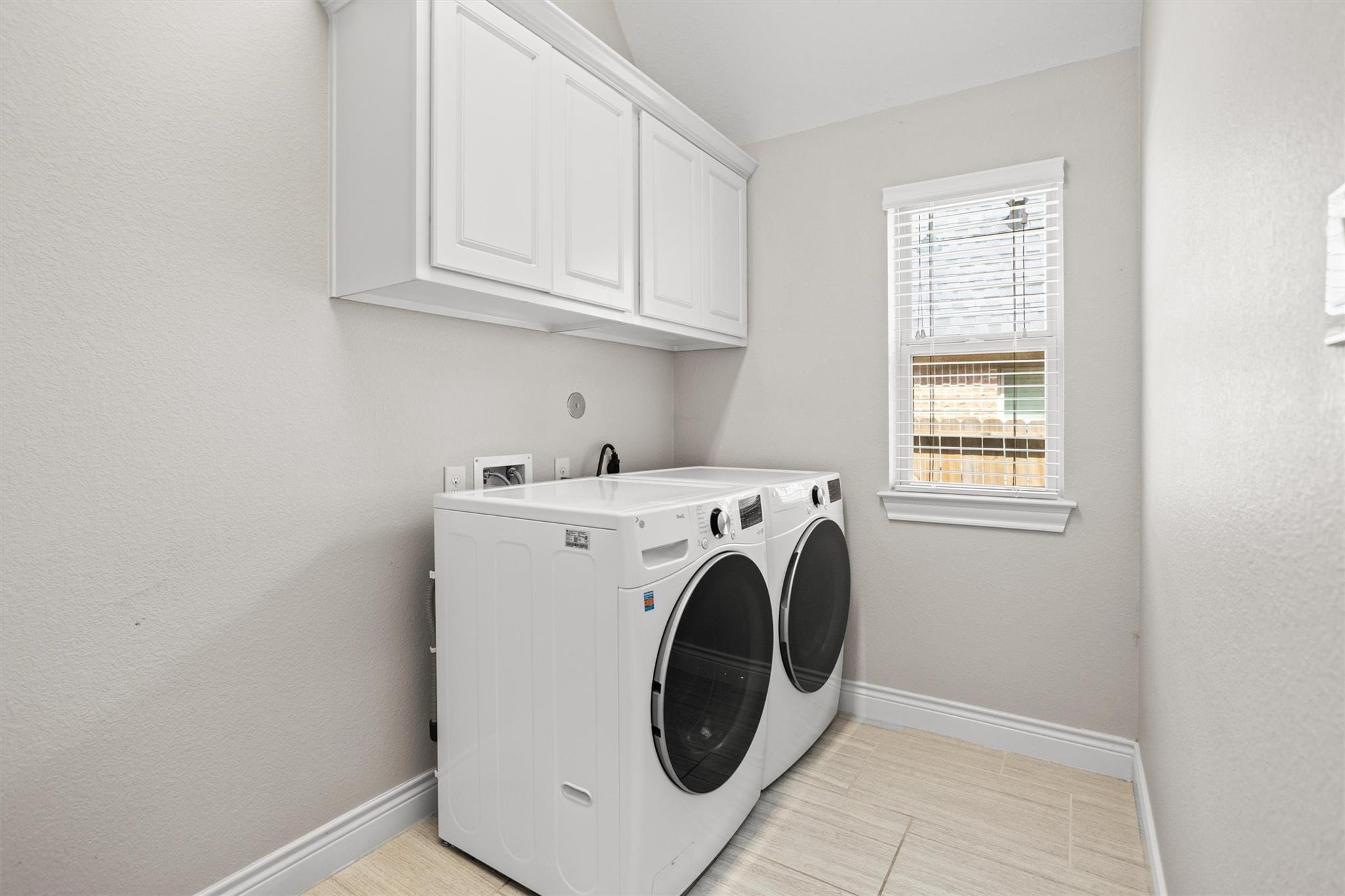 Laundry room with front-loading washer and dryer beneath white cabinets, beside a window with blinds.
