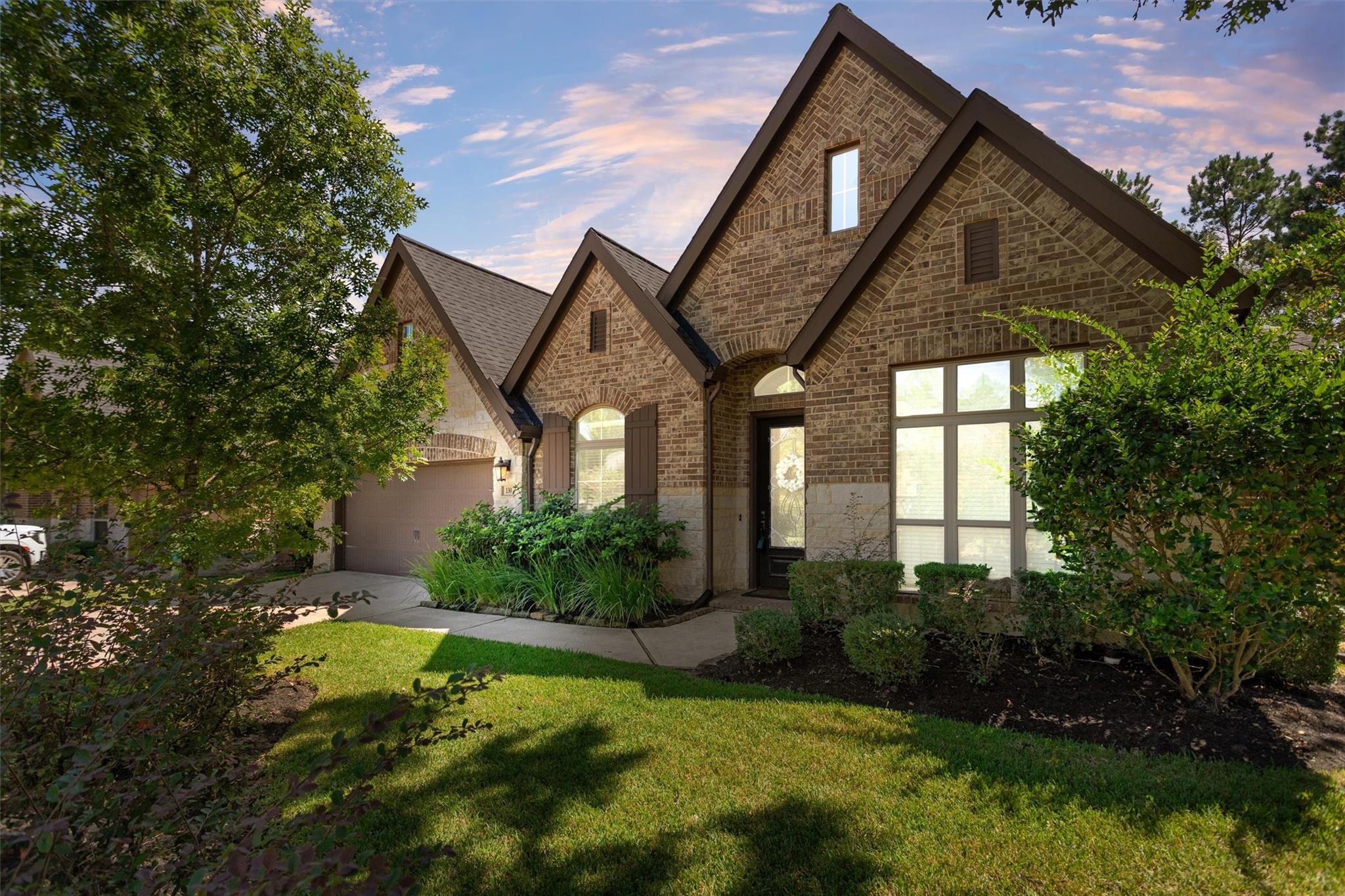 Brick house with steep gables, a garage, landscaped shrubs, and a sunny front lawn.