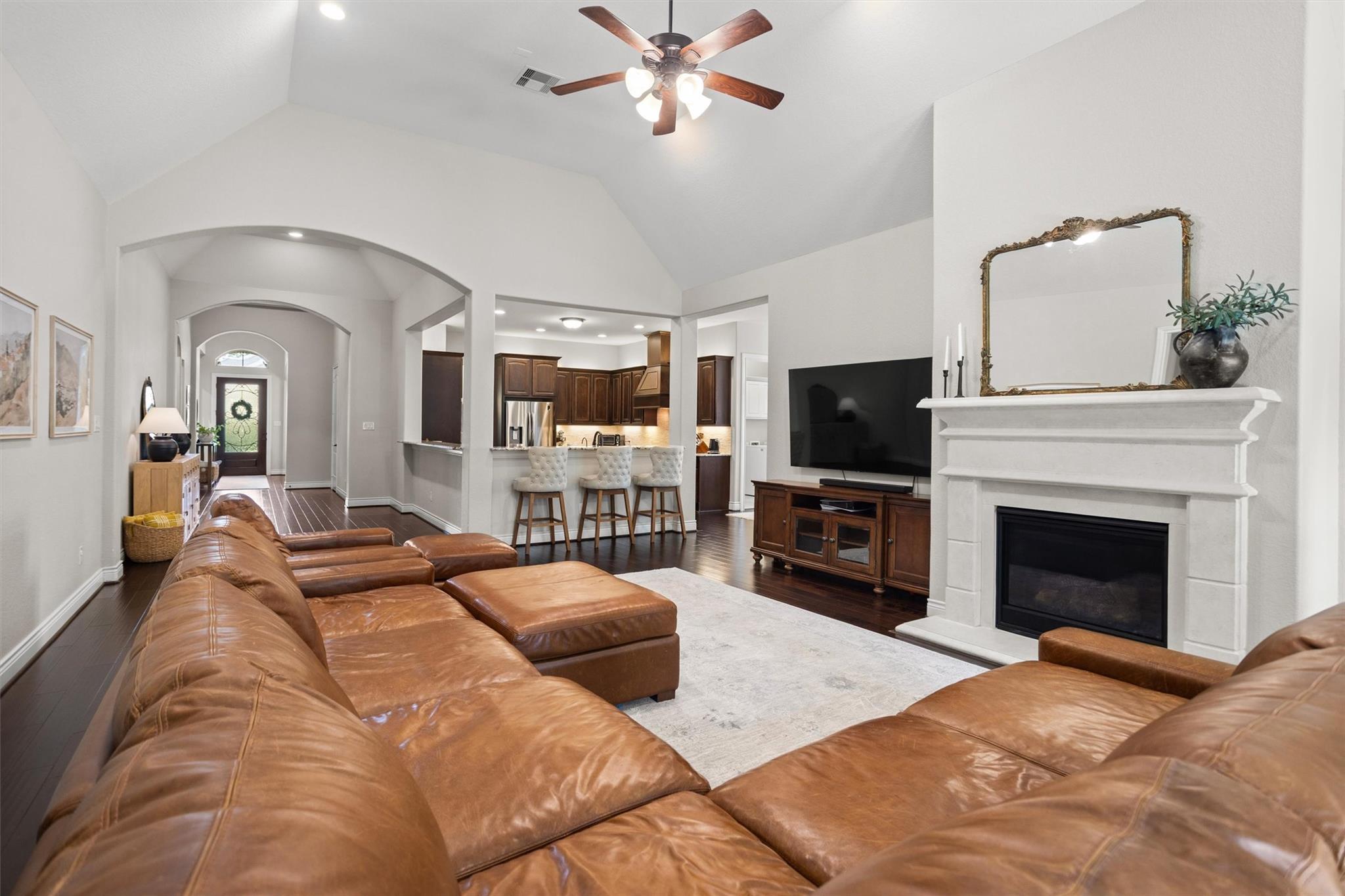 Living room with a brown leather sectional, white fireplace, vaulted ceiling, and open kitchen.
