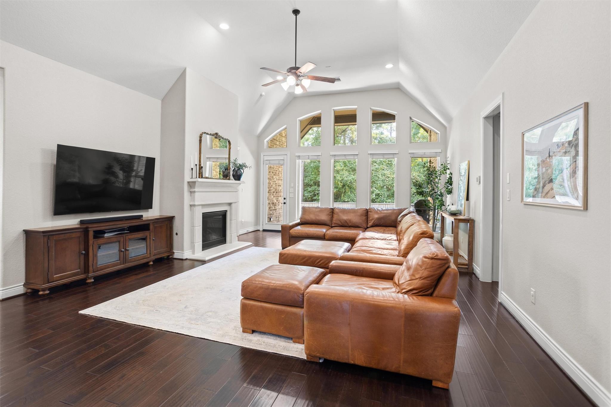 Furnished living room with vaulted ceiling, tall windows, and a brown leather sectional facing a fireplace and television.