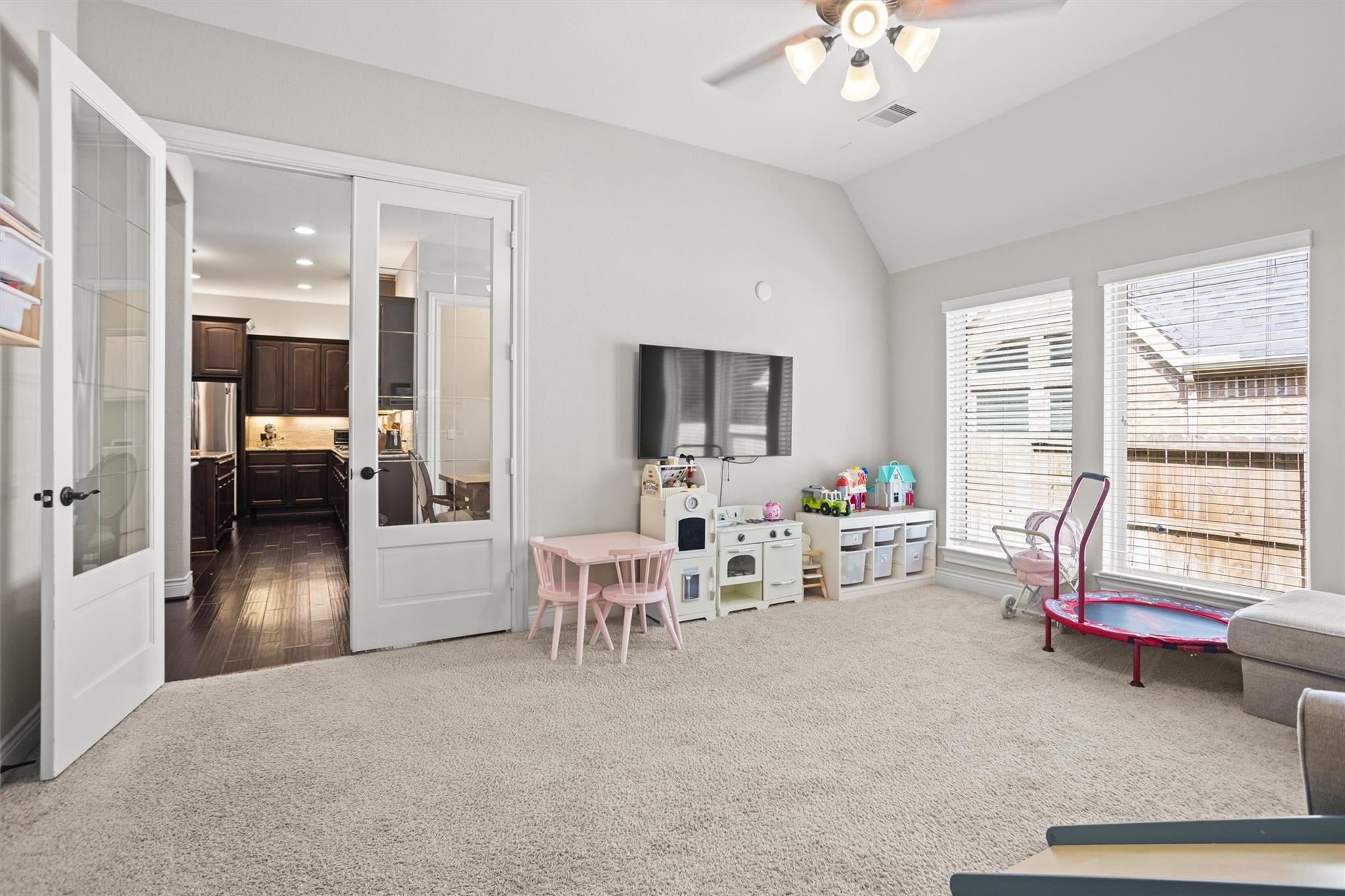 Playroom with vaulted ceiling, children’s toys, wall-mounted TV, and French doors opening toward the kitchen.