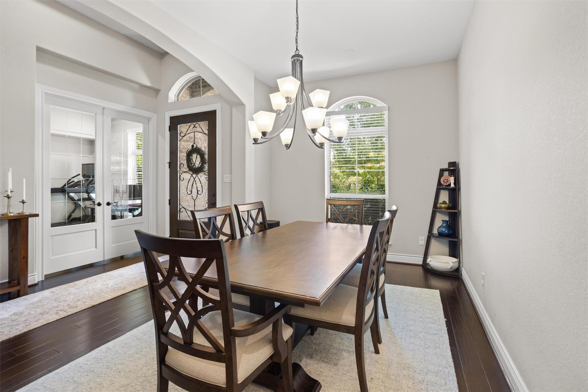 Dining room with a long wooden table, chandelier, arched window, and glass-paneled doors.