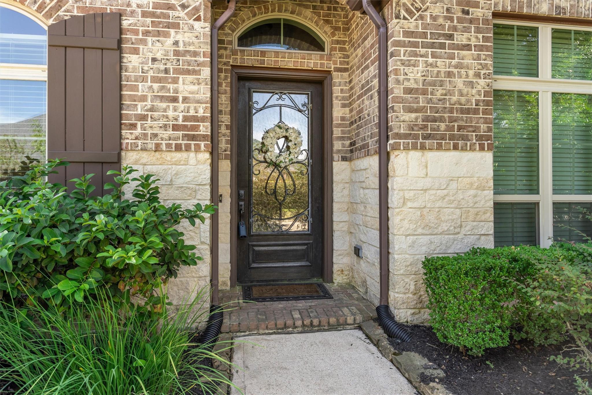Brick and stone home entrance with a dark glass door, floral wreath, and landscaped walkway.