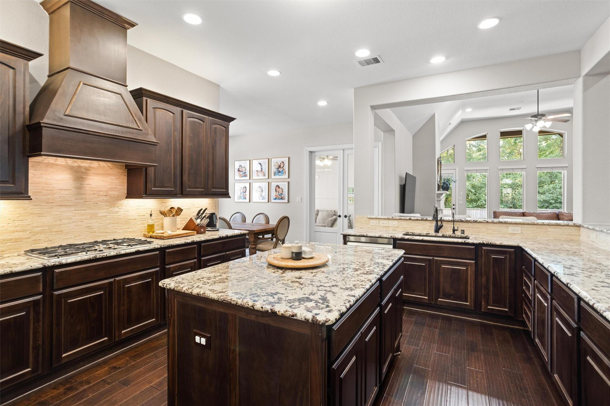Kitchen with dark wood cabinets, granite countertops, a large island, and an open view into a bright living area.