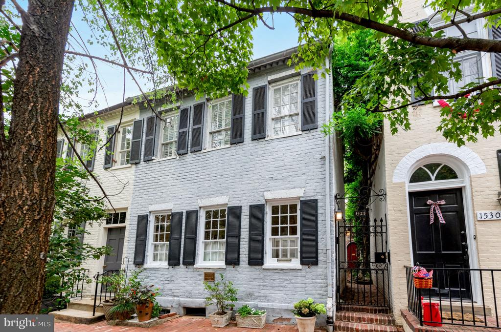Gray brick Georgetown townhouse with black shutters, leafy tree canopy, and an ornate gated entrance.