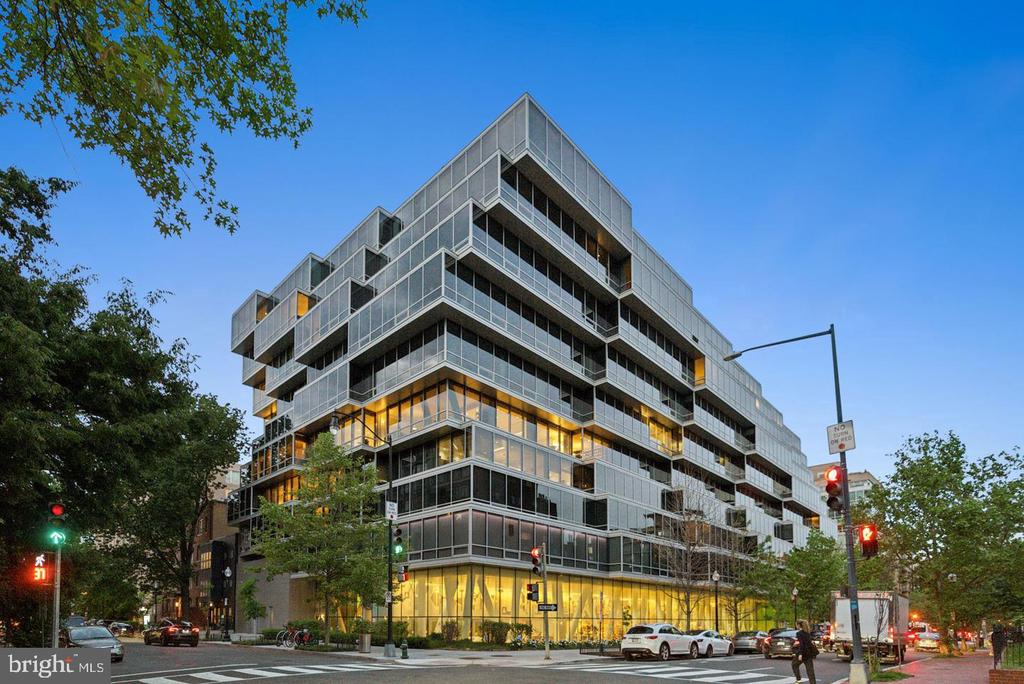 Modern glass-fronted multistory building at a city intersection, with projecting balconies and illuminated ground-floor windows.