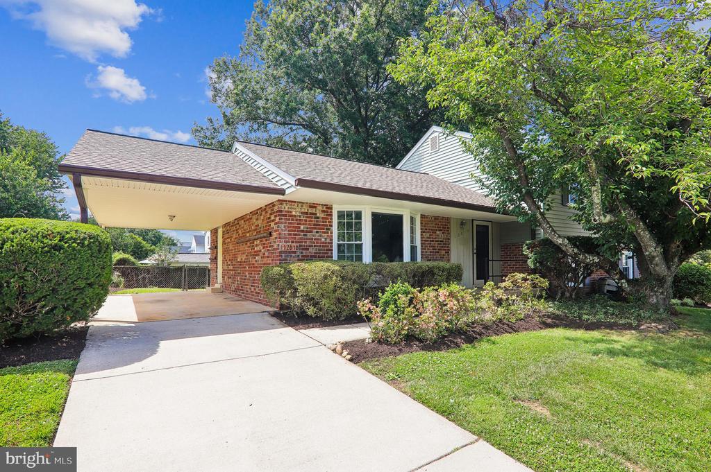 Brick house with a covered carport, broad driveway, trimmed shrubs, and mature trees.