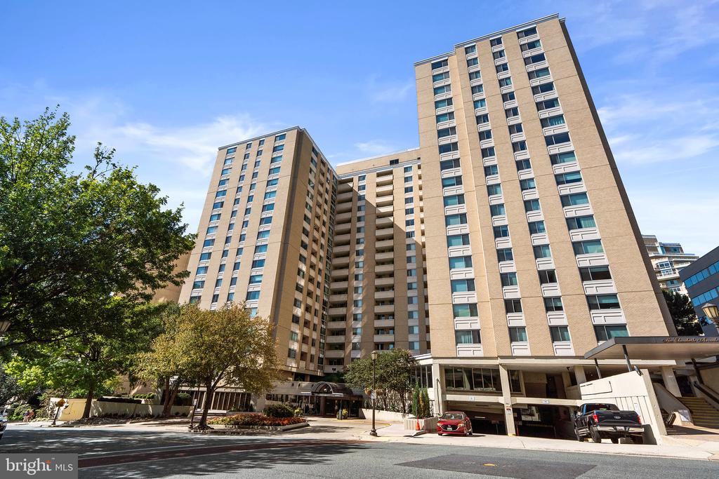 Multi-story tan condominium building with rows of windows and balconies, viewed from the street.