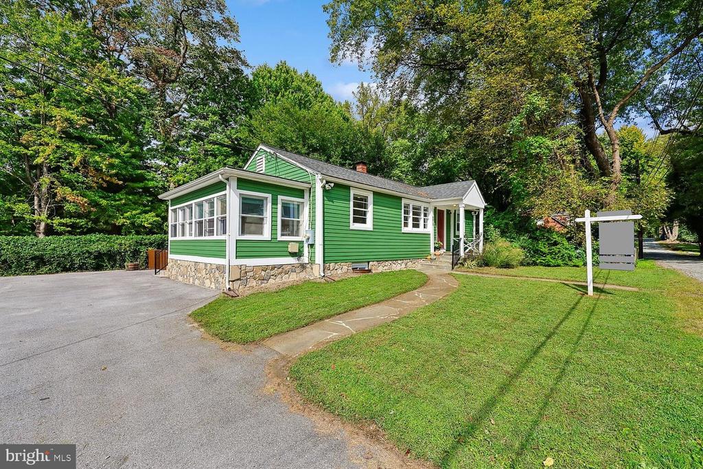Green single-story house with a stone foundation, enclosed porch, and paved driveway beneath mature trees.