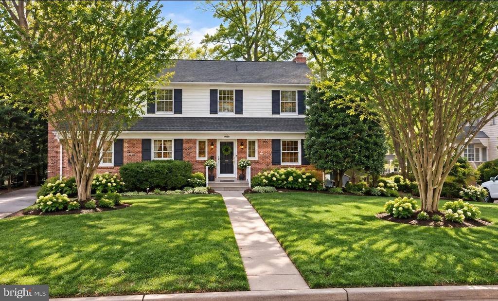 Two-story brick and white-sided house with black shutters, mature trees, and a landscaped front lawn.