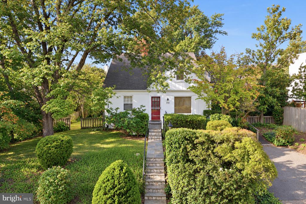 White Cape Cod-style house with a red front door, stone walkway, and mature trees.