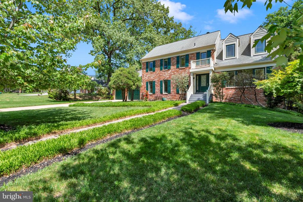 Brick house with dormer windows and a white balcony behind a shady front lawn.