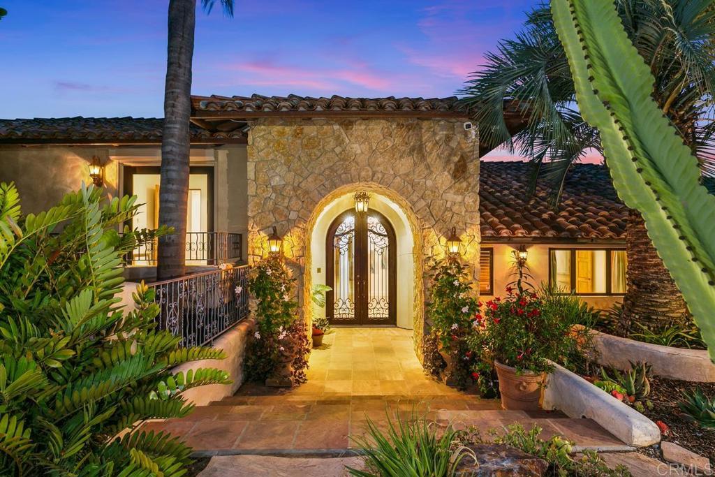 Rancho Santa Fe home with a warmly lit stone archway, wrought-iron doors, and tropical landscaping at dusk.