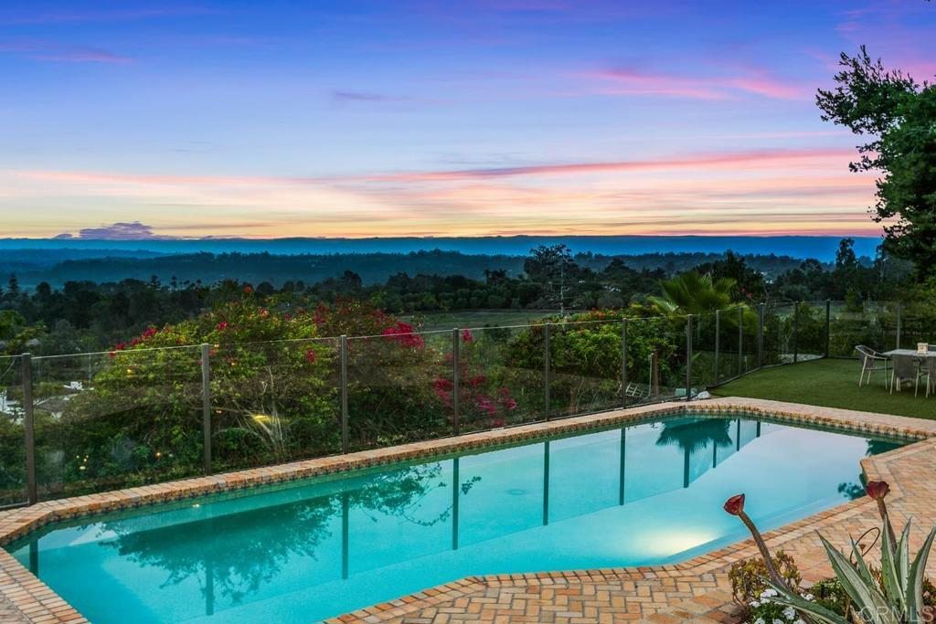 Swimming pool overlooking wooded hills at sunset, bordered by a glass fence and brick patio.