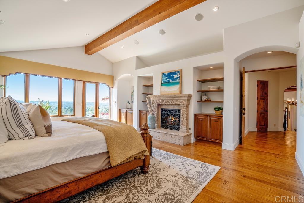 Bedroom with a carved stone fireplace, exposed wood beam, and large windows overlooking a distant landscape.
