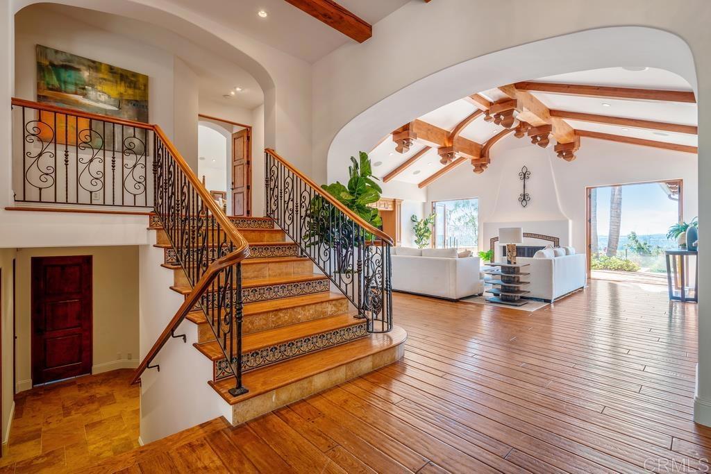 Open living room and entry with a sweeping wood staircase, decorative iron railings, and exposed ceiling beams.