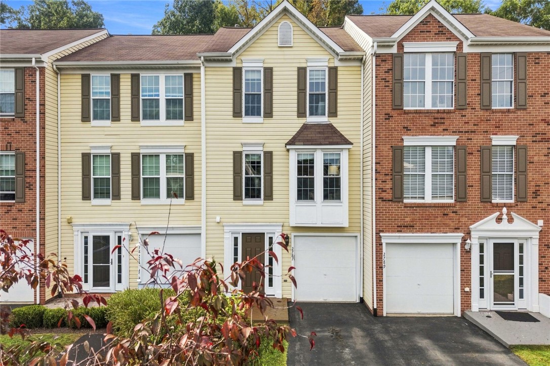 Yellow-sided townhouse with dark shutters, a white garage door, and neighboring brick homes.