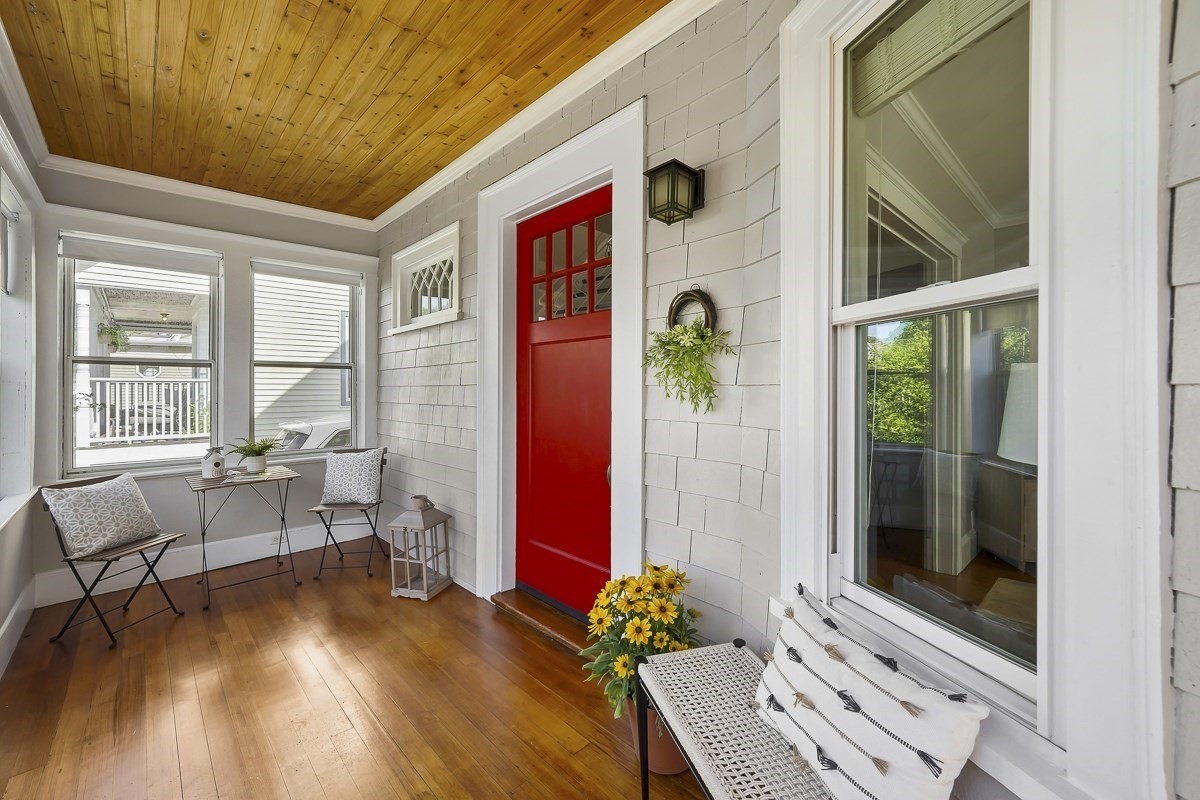 Enclosed front porch with a bright red door, wood ceiling, hardwood floor and window-side seating.